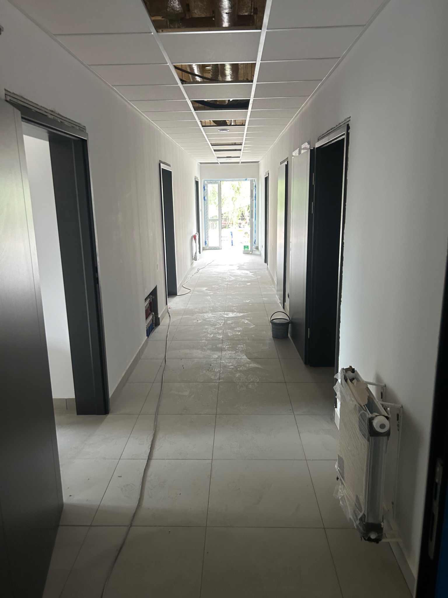 Korytarz biurowy w trakcie remontu: widoczne przygotowane ściany do malowania, częściowo zamontowany sufit podwieszany, zabezpieczony grzejnik i wiadro na podłodze.