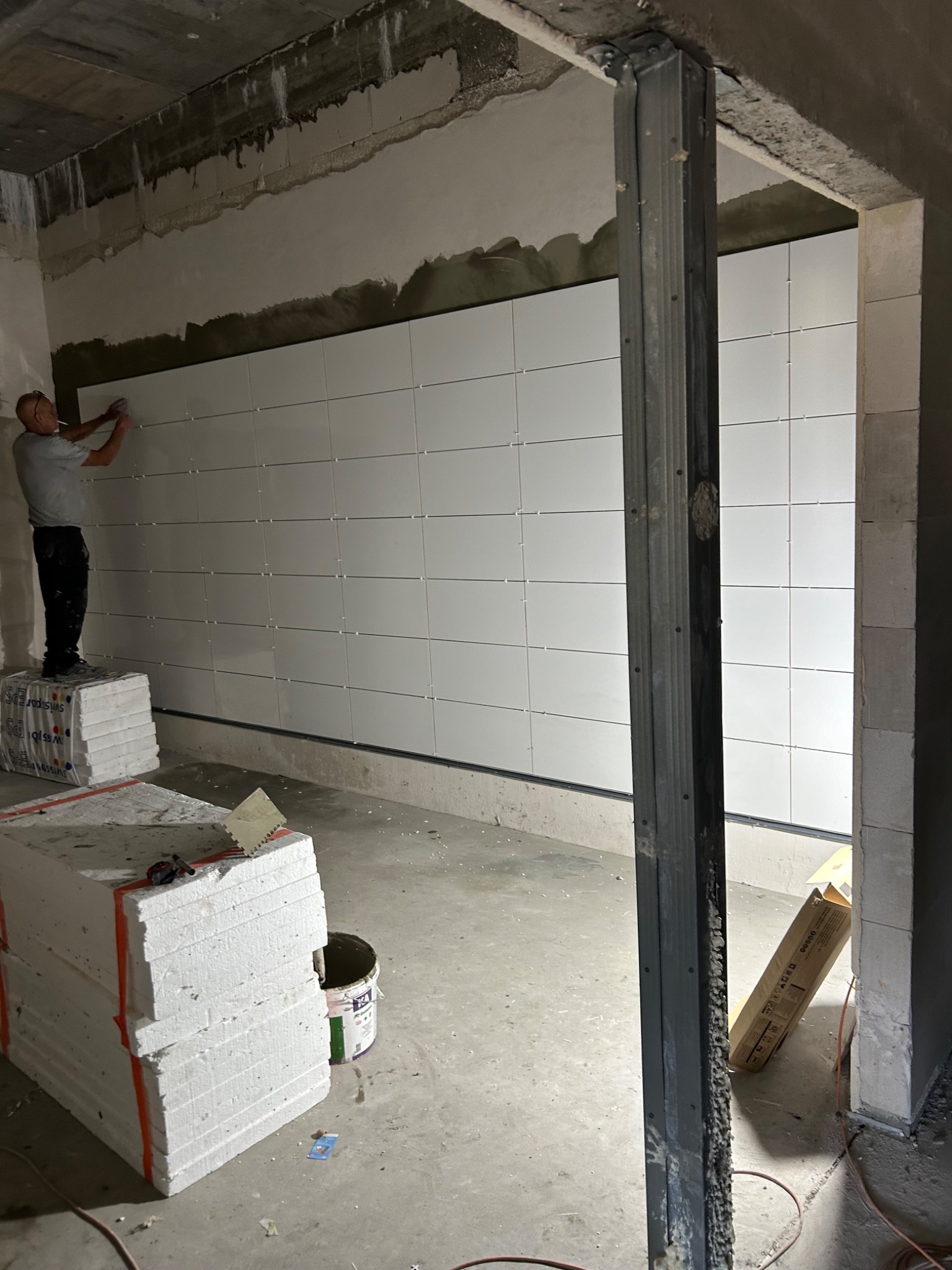 Pracownik na podwyższeniu układa białe płytki na ścianie w surowym wnętrzu, widoczne materiały budowlane i narzędzia.