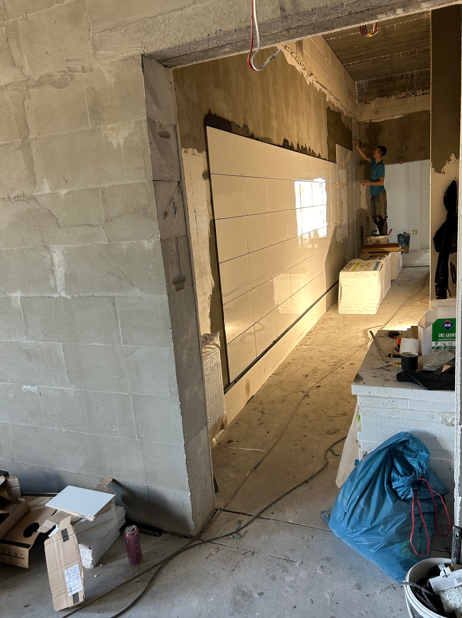Wnętrze w trakcie remontu: pracownik na podwyższeniu układa białe płytki na ścianie, widoczne surowe ściany z betonu komórkowego, materiały budowlane i narzędzia, niebieski worek na odpady.