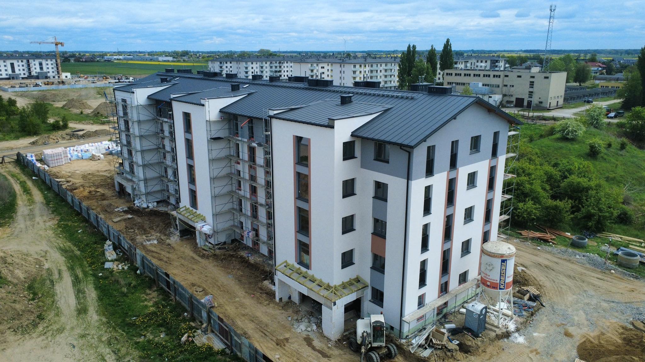 Wysoki budynek mieszkalny w trakcie budowy, z widocznymi rusztowaniami. Uwagę zwraca zbiornik z zaprawą na placu budowy i częściowo wykończona elewacja w tonacji bieli i szarości.