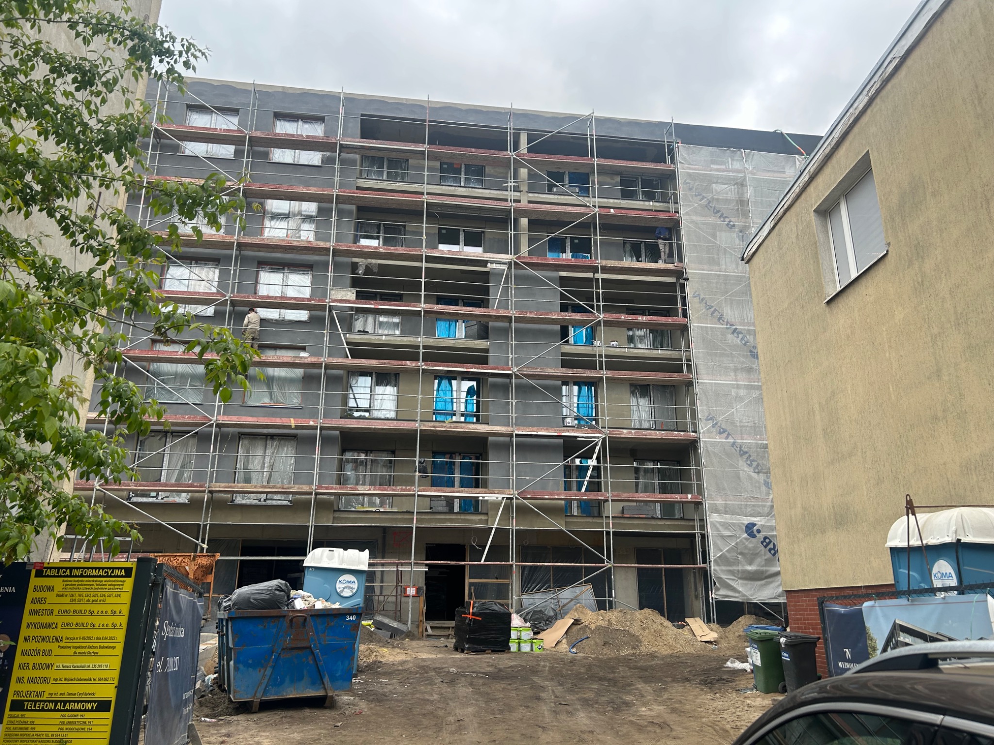 Wielopiętrowy budynek w trakcie prac elewacyjnych. Widoczne rusztowania, częściowo ocieplona ściana, okna zabezpieczone folią. Prace w toku, widoczny pracownik.