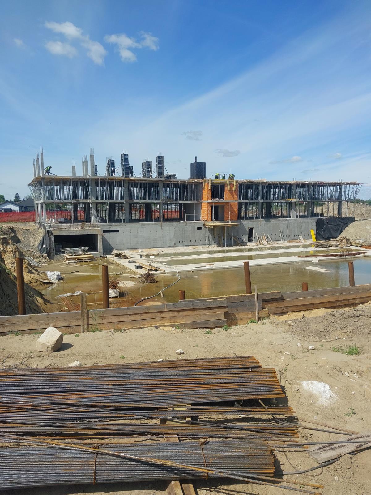 Budowa dwukondygnacyjnego budynku w stanie surowym otwartym z widocznym zbrojeniem i elementami betonowymi, na tle błękitnego nieba z chmurami.