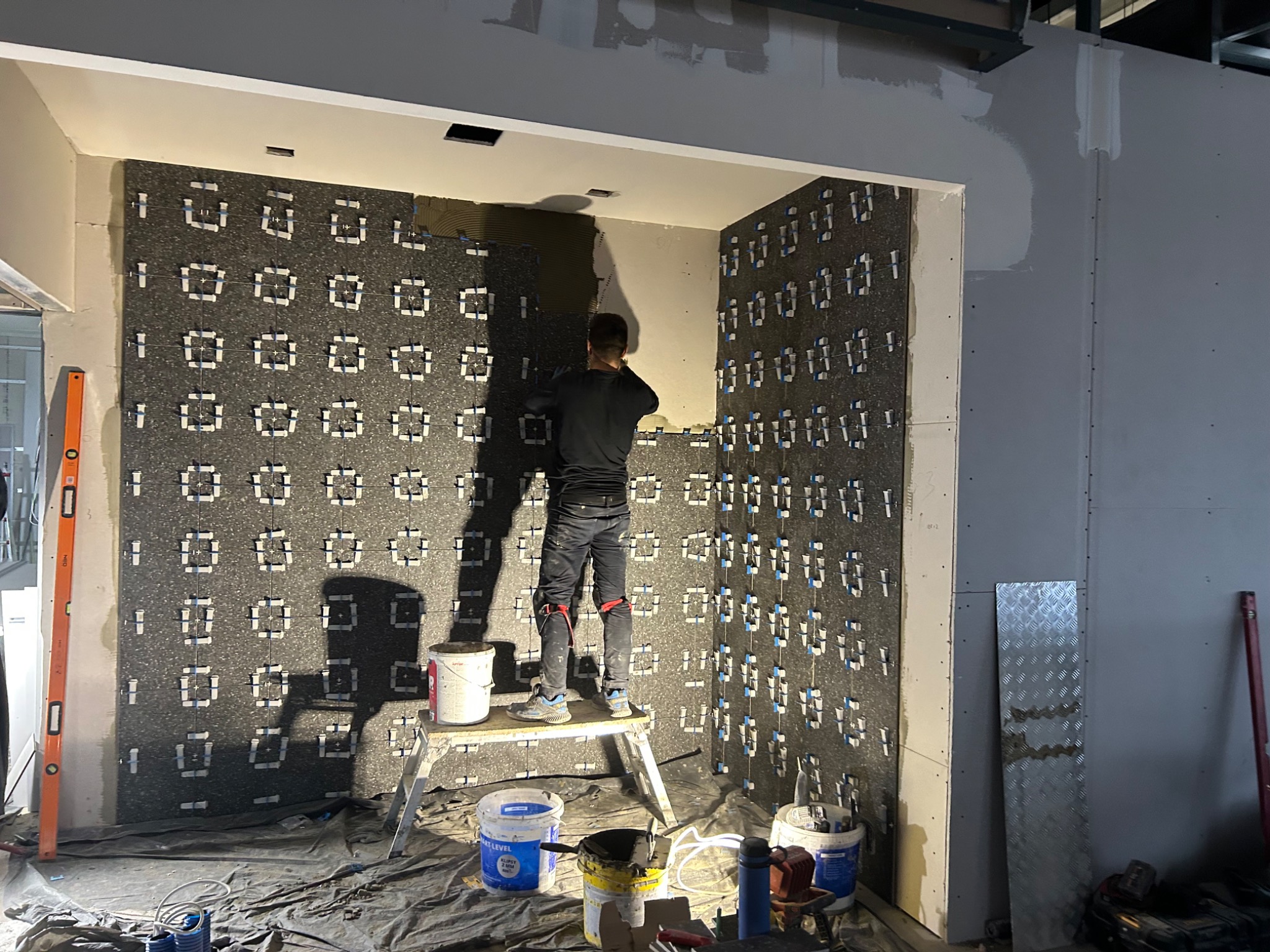 Pracownik na podeście aplikuje zaprawę na ścianę z zamocowaną izolacją akustyczną. Widoczne narzędzia, poziomica i wiaderka z zaprawą. Wnętrze w trakcie prac wykończeniowych.