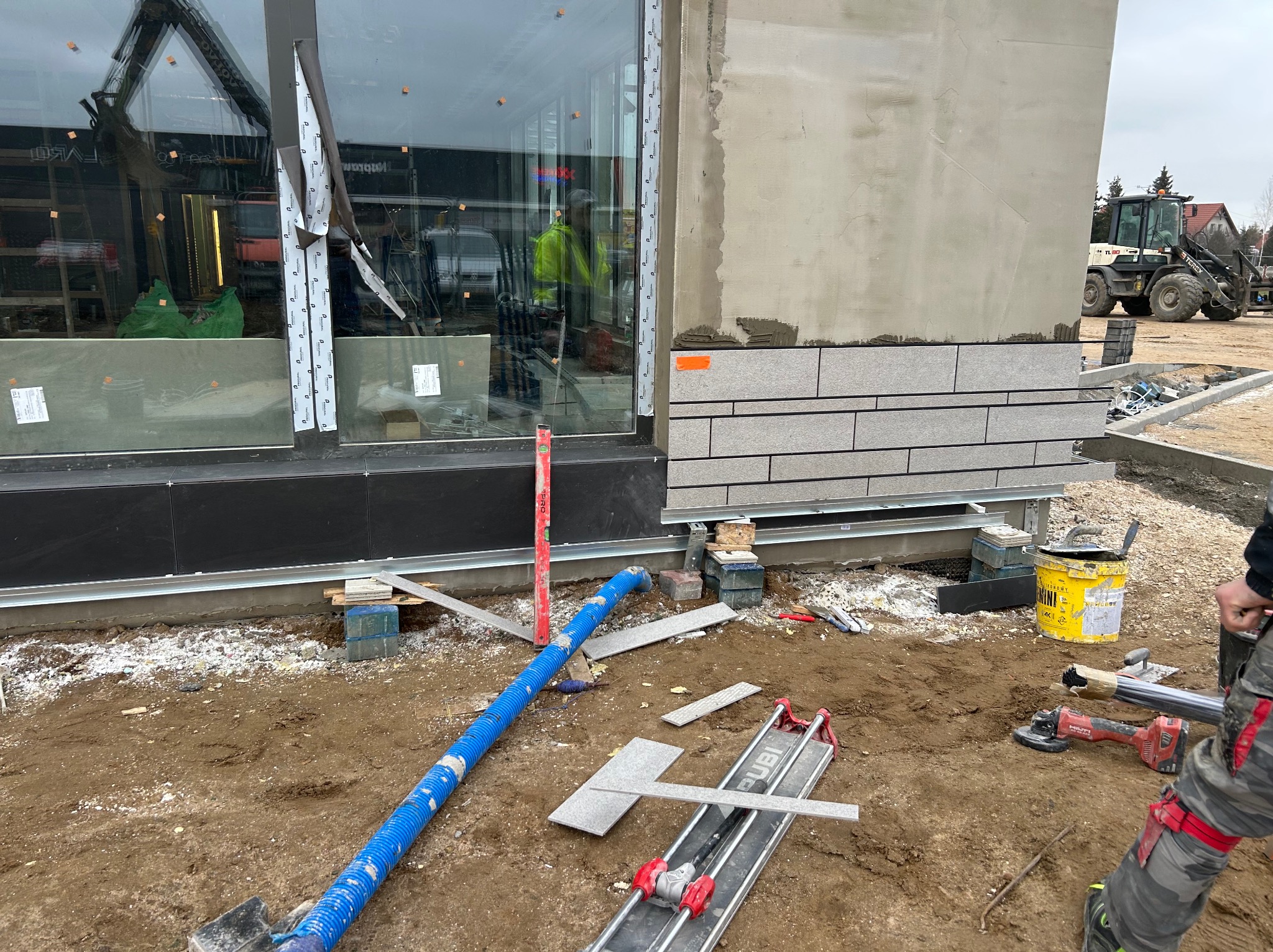 Układanie szarej, prostokątnej okładziny elewacyjnej na budynku. Widoczne narzędzia glazurnicze, poziomica i fragment koparki w tle. Prace w toku.