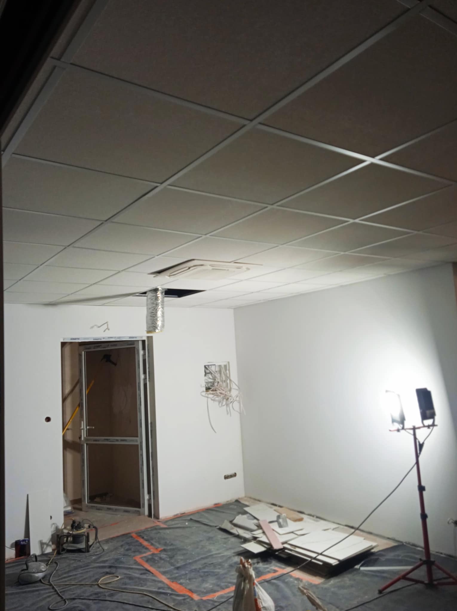 Montaż sufitu podwieszanego kasetonowego w biurze, widoczne kable, lampa budowlana i przygotowane płyty. Prace wykończeniowe w trakcie realizacji.