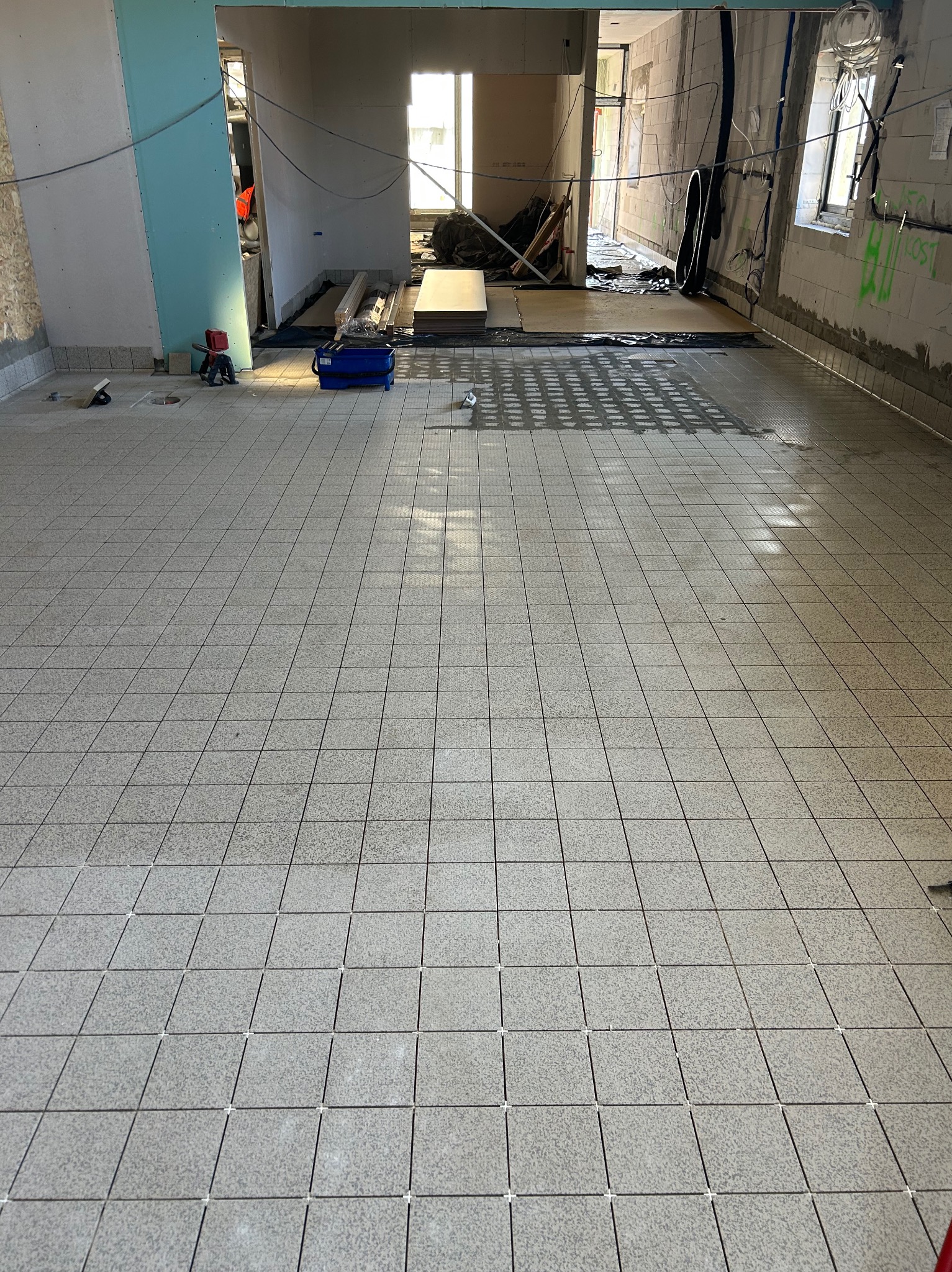 Ułożona, jasnoszara podłoga z kwadratowych płytek w trakcie remontu. Widoczne krzyżyki dystansowe, narzędzia i materiały budowlane w tle. Surowy stan ścian.