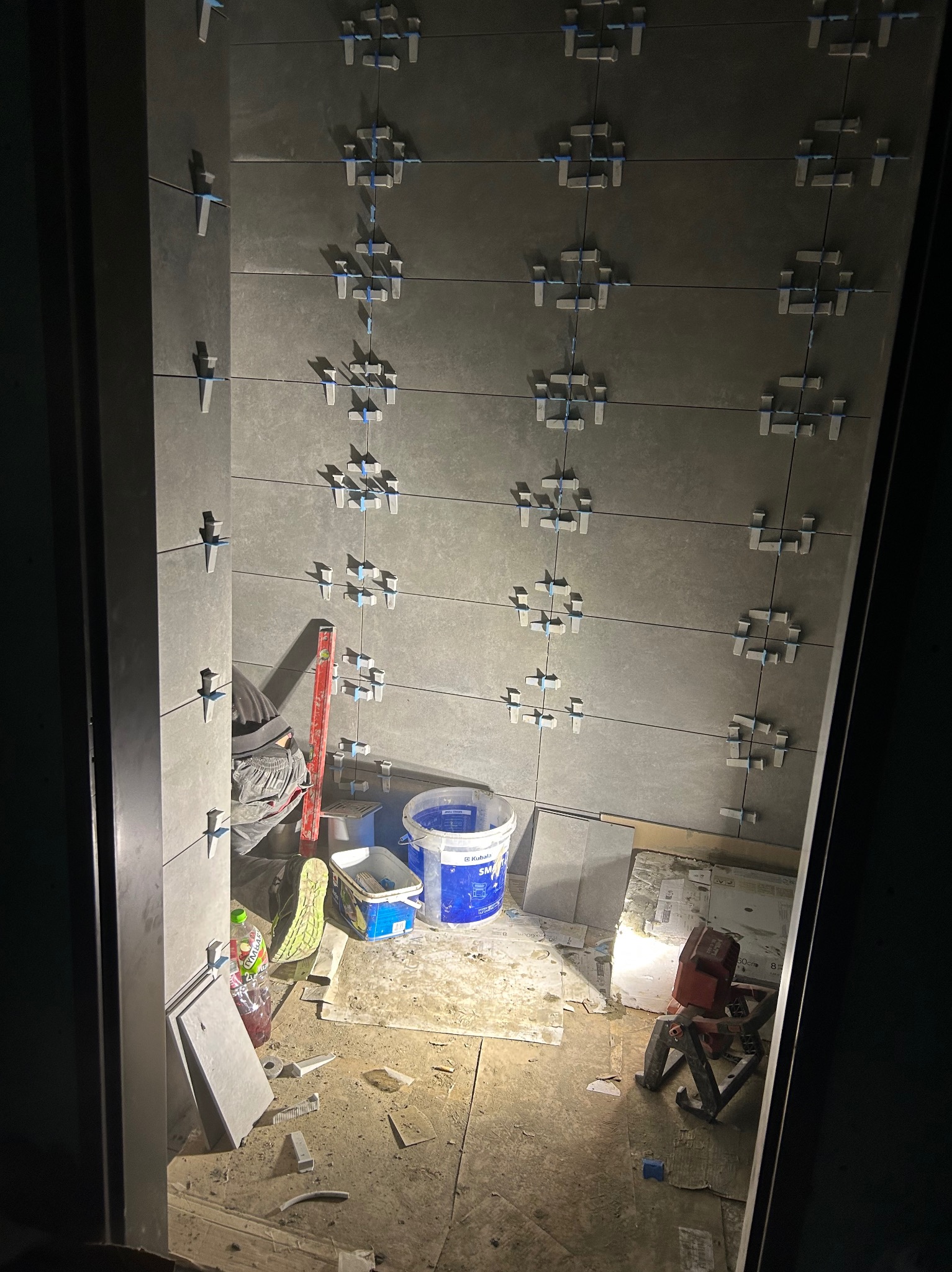 Układanie szarych płytek na ścianie z użyciem krzyżyków dystansowych, widoczne narzędzia pracy w Mławie. Praca w toku, oświetlenie robocze.
