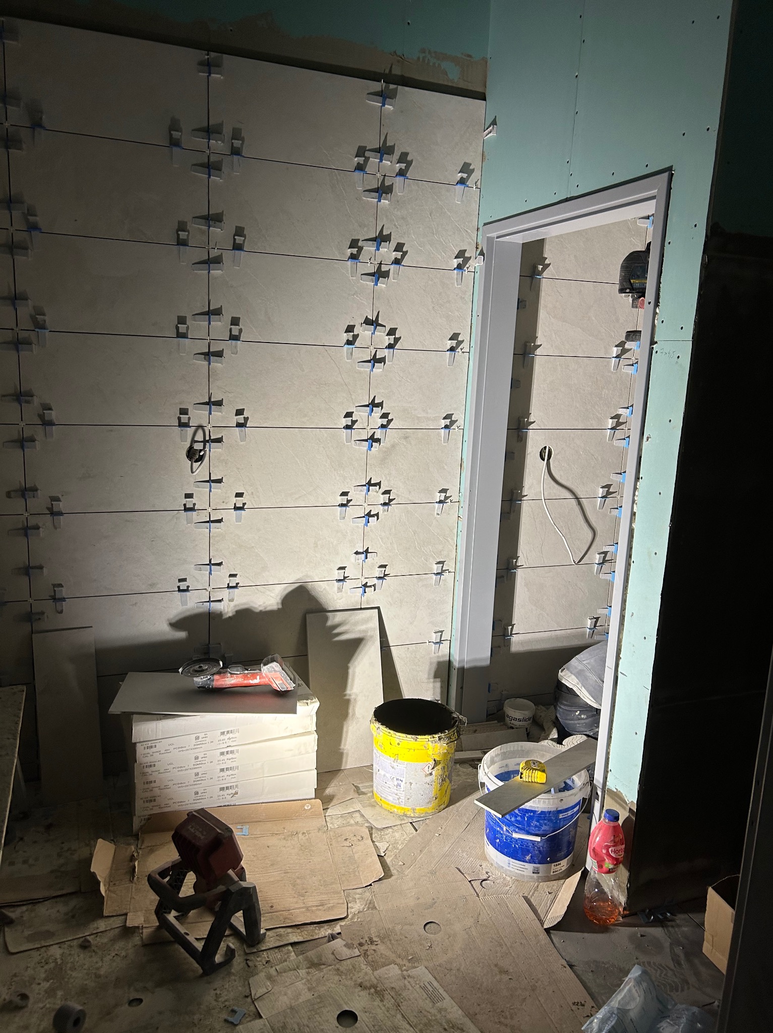 Ściana z jasnymi płytkami, krzyżyki dystansowe, poziomica laserowa, wiadro z klejem. W tle widoczne płyty gipsowo-kartonowe i ościeżnica drzwi.