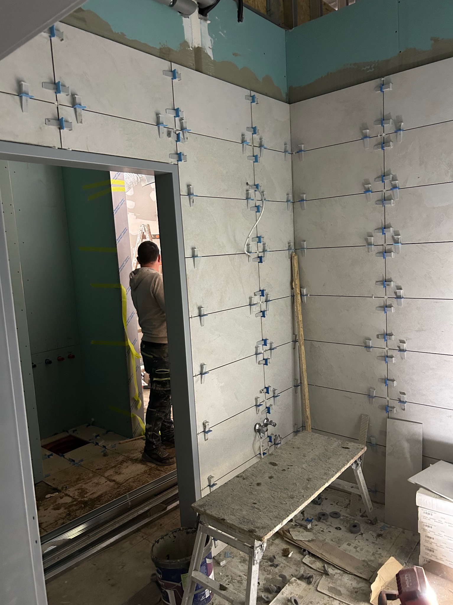 Układanie płytek w łazience z użyciem systemu poziomowania. Widoczny glazurnik w trakcie pracy, płytki na ścianach i podłodze, narzędzia i materiały budowlane.