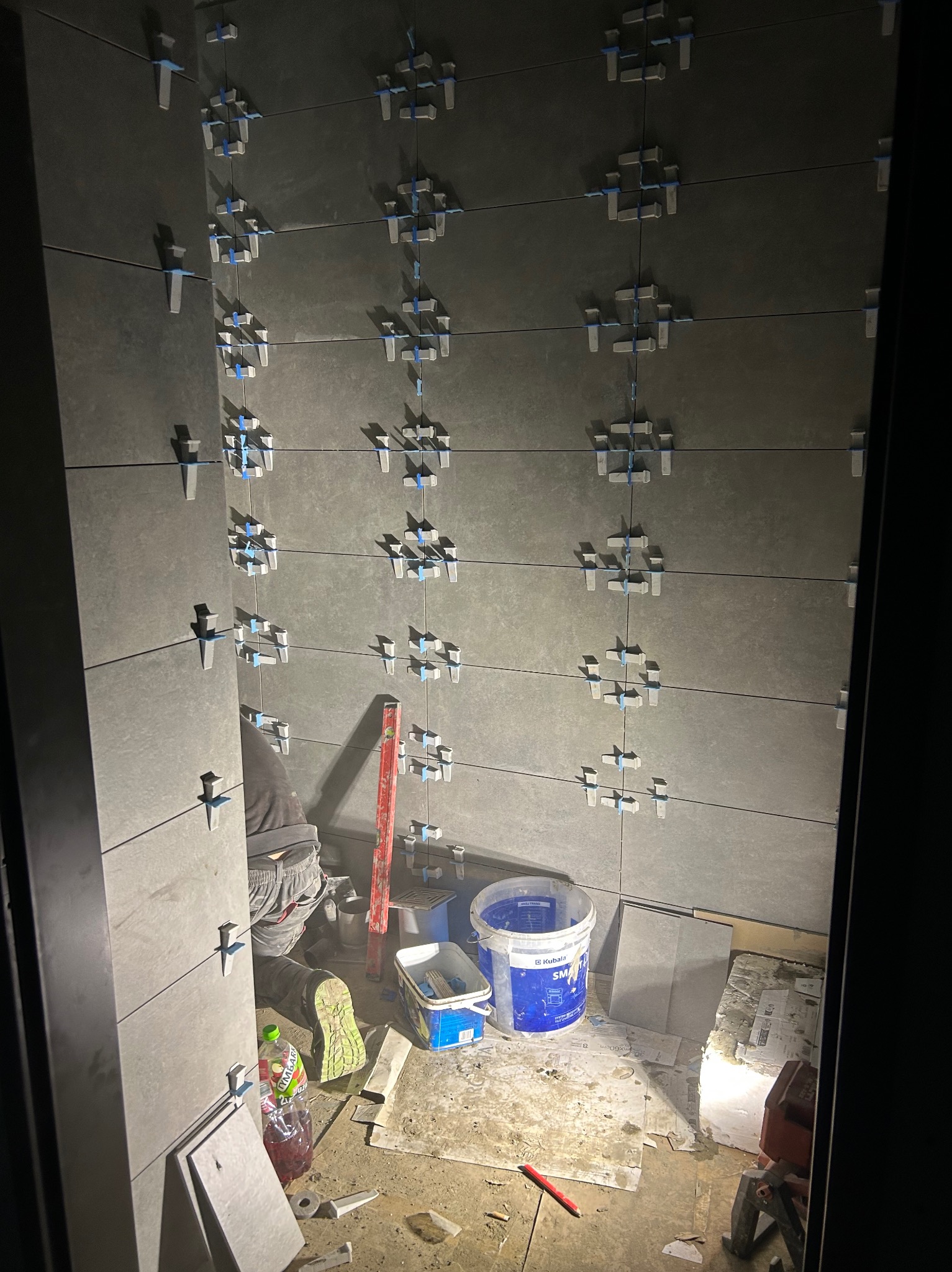 Układanie szarych płytek na ścianie z użyciem krzyżyków dystansowych. Widoczne narzędzia glazurnicze i częściowo sylwetka glazurnika w trakcie pracy. Ujęcie z dołu.