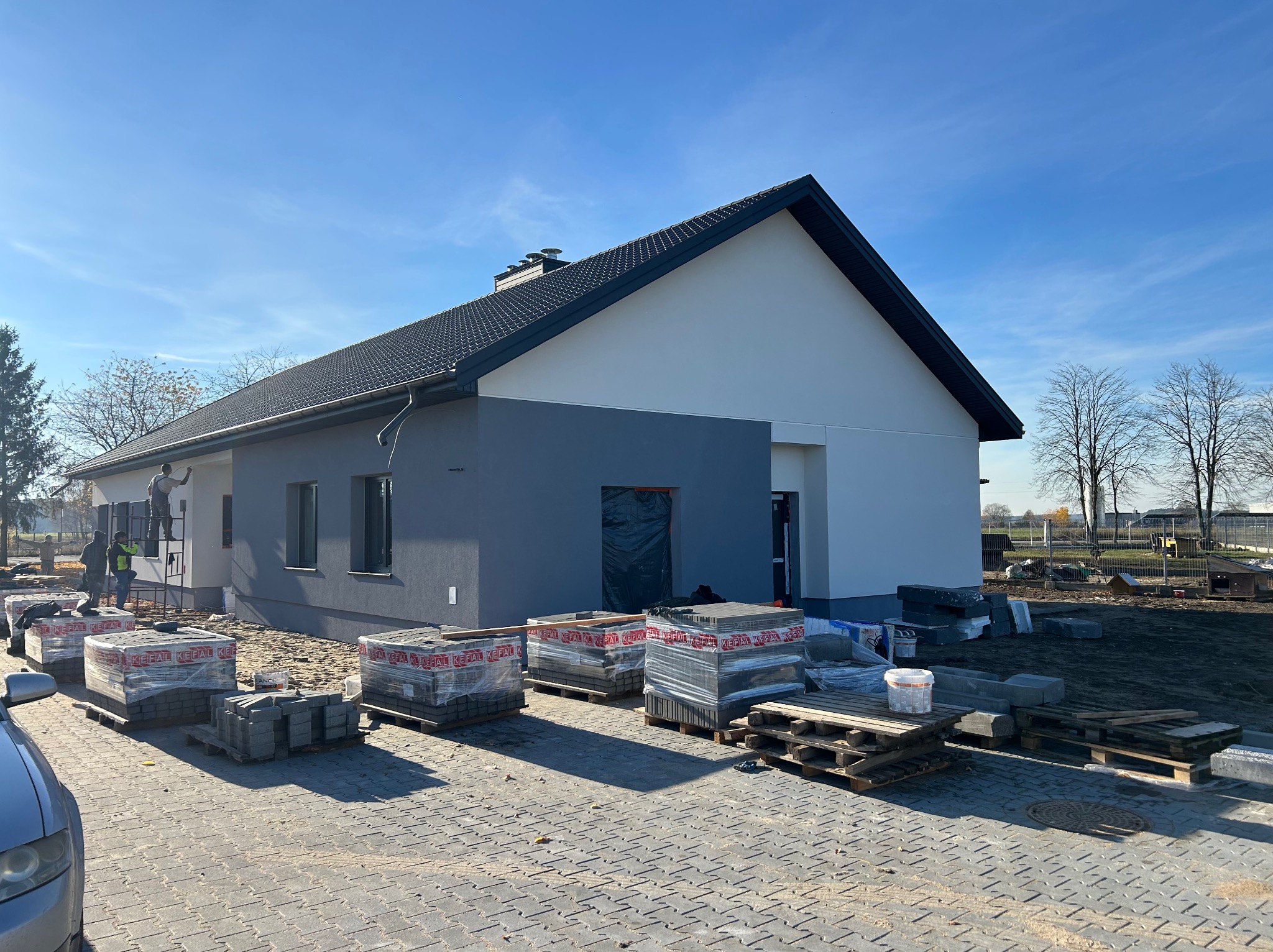 Nowoczesny dom jednorodzinny z szarą i białą elewacją, czarnym dachem i ułożoną kostką brukową. Na pierwszym planie palety z kostką. Pracownik na drabinie przy elewacji.