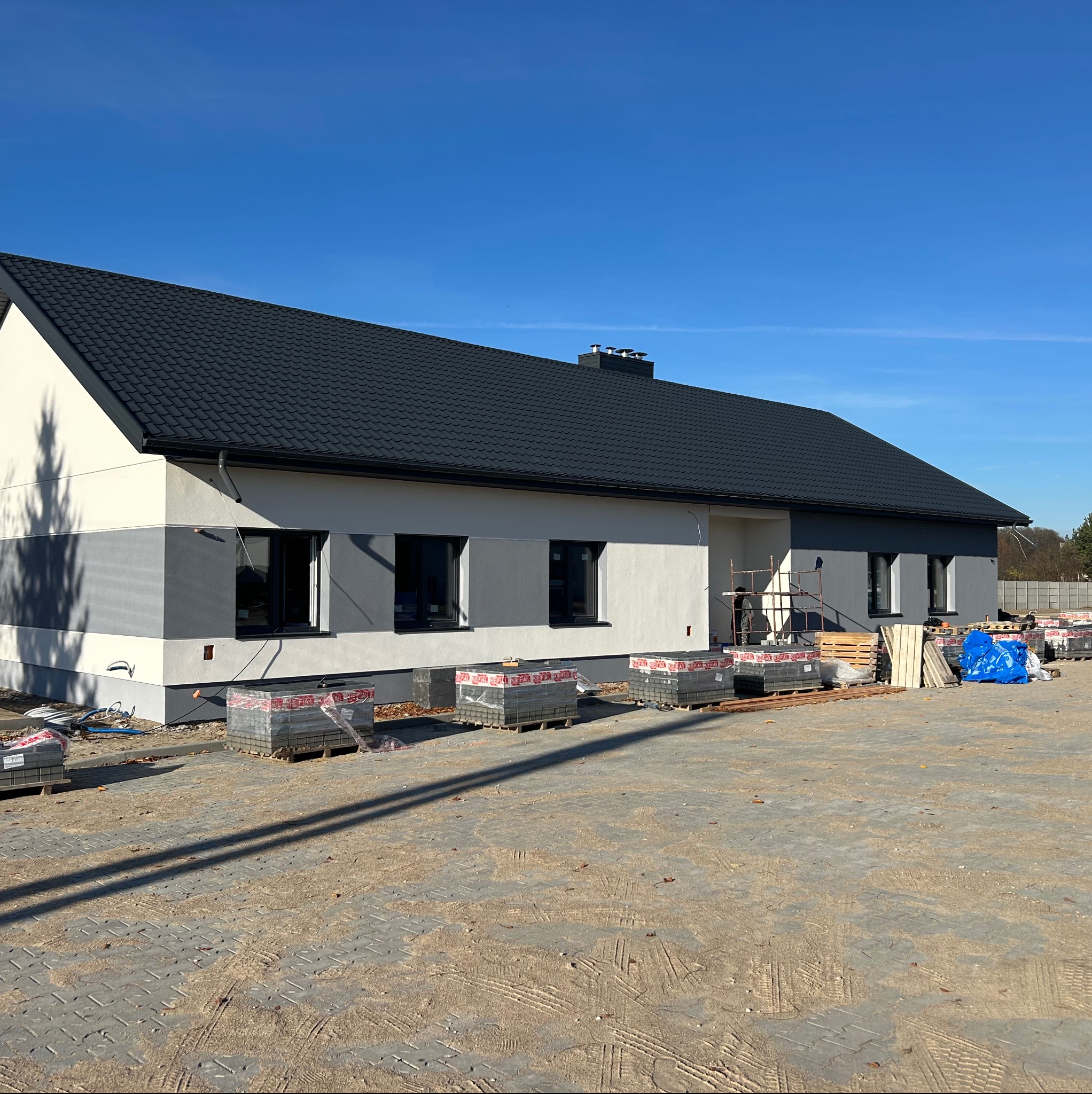 Nowoczesny dom jednorodzinny z szarą elewacją i ciemnym dachem, kostka brukowa na podjeździe, materiały budowlane przed budynkiem w słoneczny dzień.