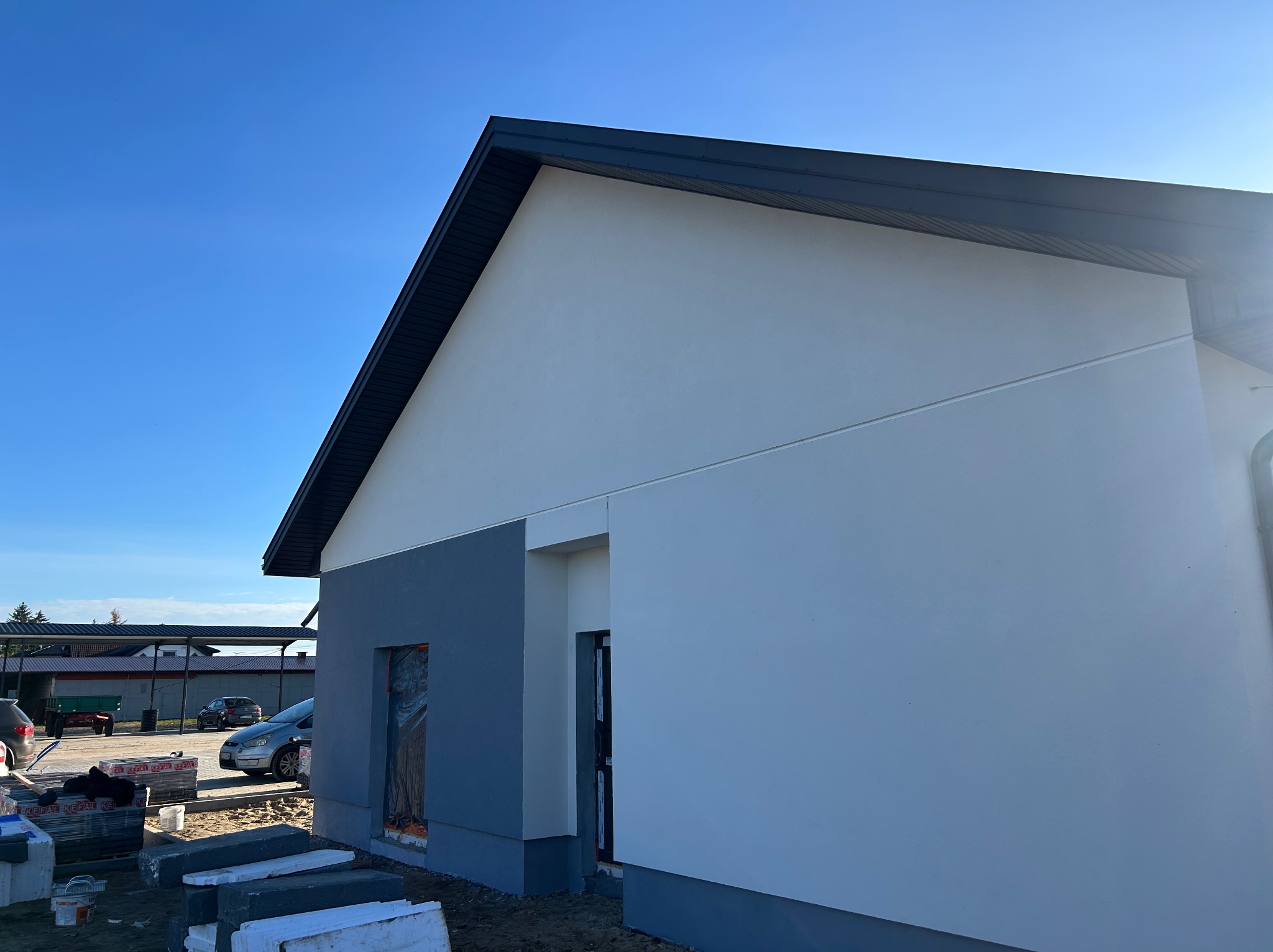 Nowoczesny dom jednorodzinny z szarą elewacją i ciemnym dachem na tle błękitnego nieba. Widoczne materiały budowlane sugerują etap wykończeniowy.