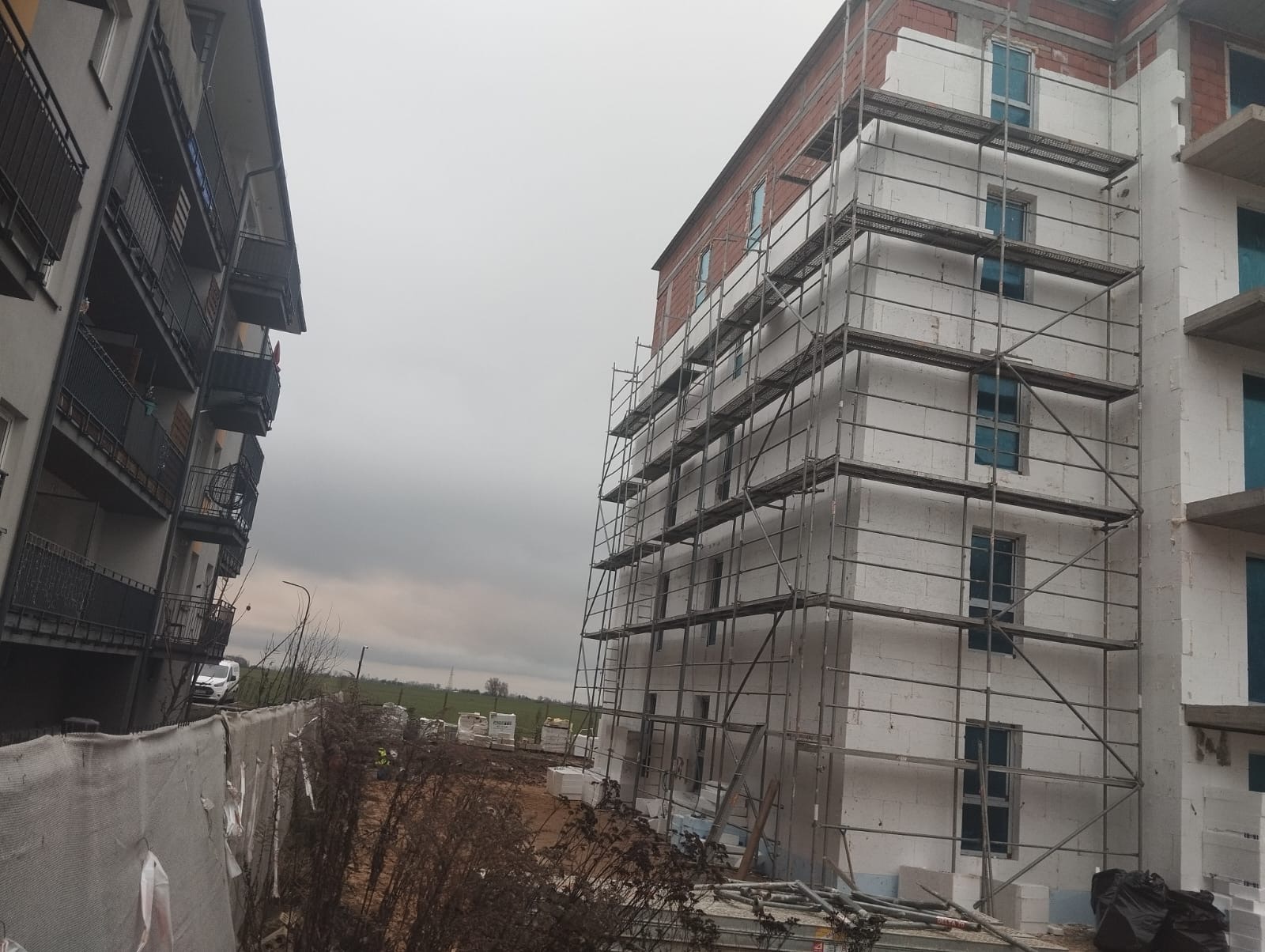 Budynek w trakcie ocieplania, z widocznymi rusztowaniami i materiałami izolacyjnymi. Częściowo ocieplona elewacja, widoczne okna. W tle inny budynek i pochmurne niebo.