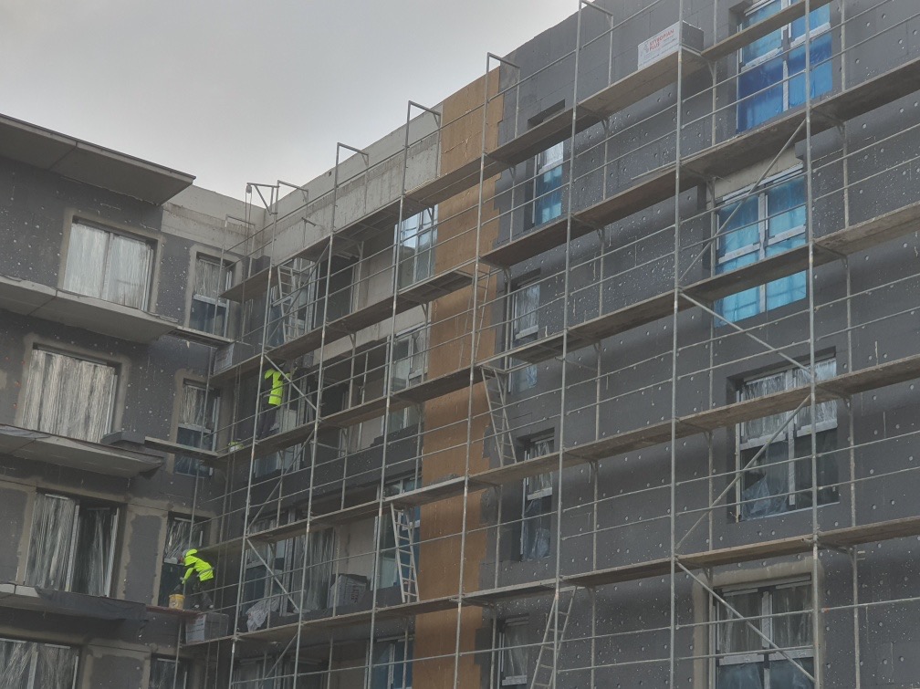 Budynek wielorodzinny w trakcie ocieplania. Widoczne rusztowania, pracownicy w odblaskowych ubraniach i fragmenty ocieplonej elewacji. Okna zabezpieczone folią.