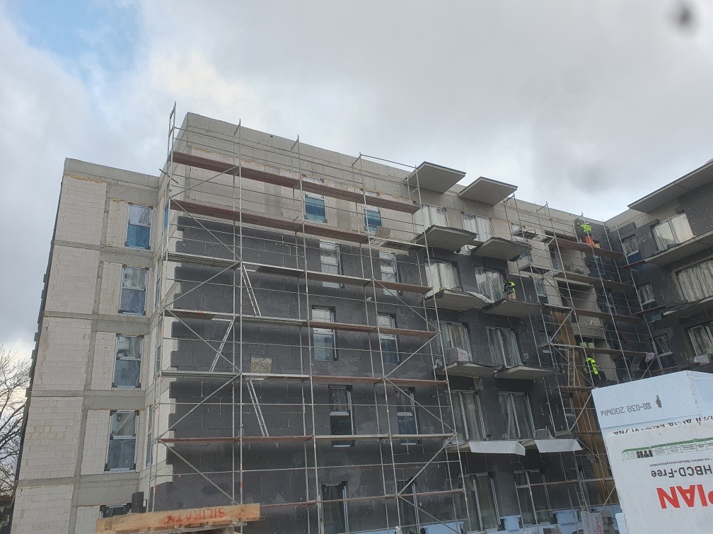 Budynek wielorodzinny w trakcie prac elewacyjnych. Widoczne rusztowania, ocieplenie ścian styropianem grafitowym i pracownicy w odblaskowych kamizelkach. Pochmurne niebo w tle.