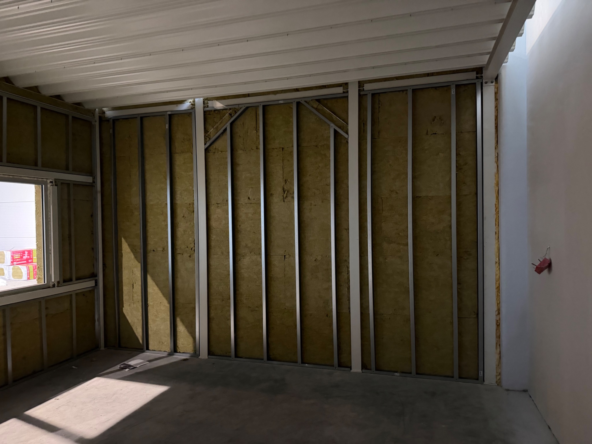 Wnętrze w trakcie prac: metalowy szkielet ściany wypełniony wełną mineralną, widoczne pakiety materiałów przy oknie, surowy beton na podłodze, przygotowanie do dalszych prac wykończeniowych.