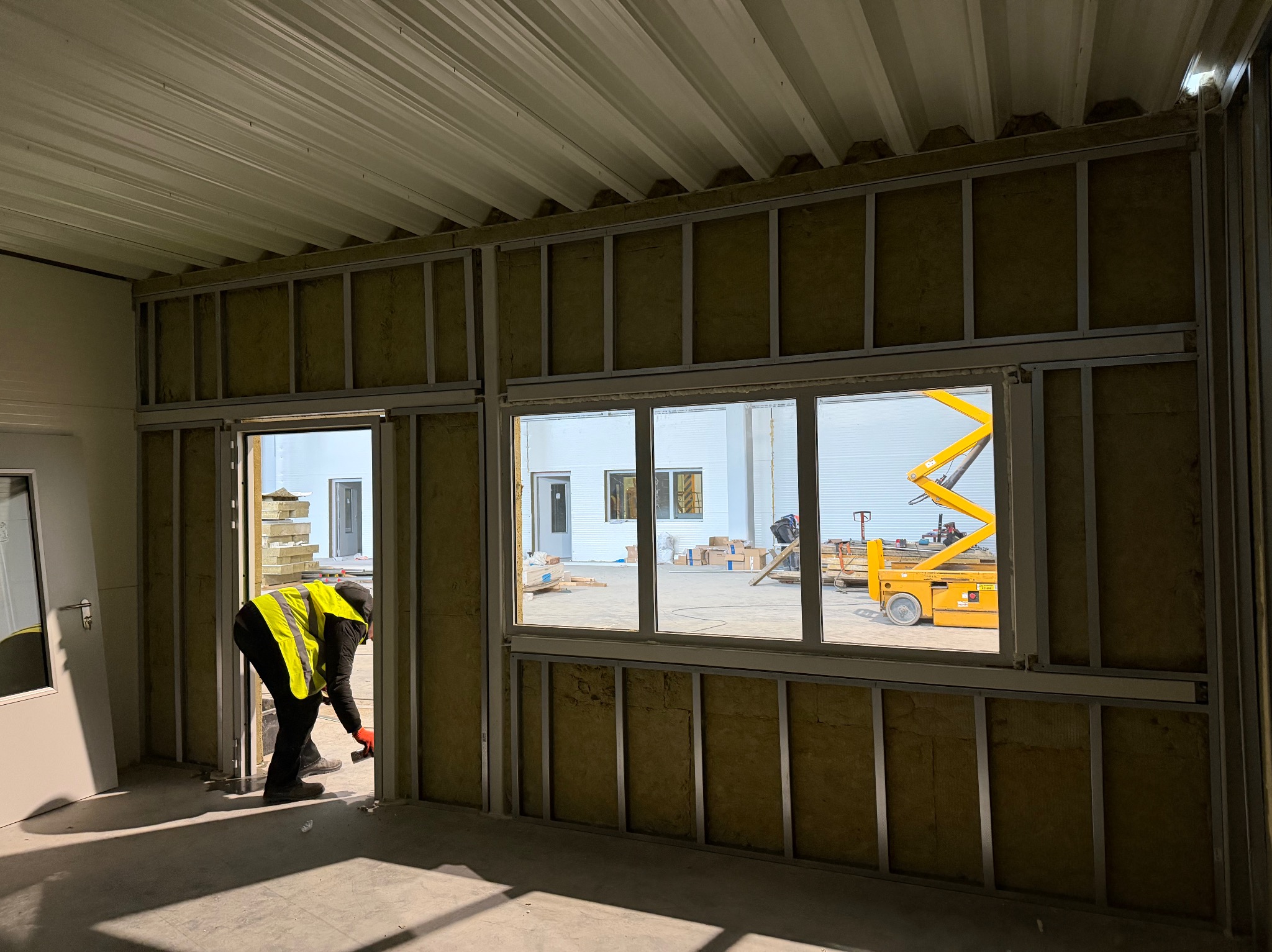 Montaż izolacji z wełny mineralnej w metalowej konstrukcji szkieletowej. Widoczny pracownik w kamizelce odblaskowej i podnośnik koszowy za oknem.