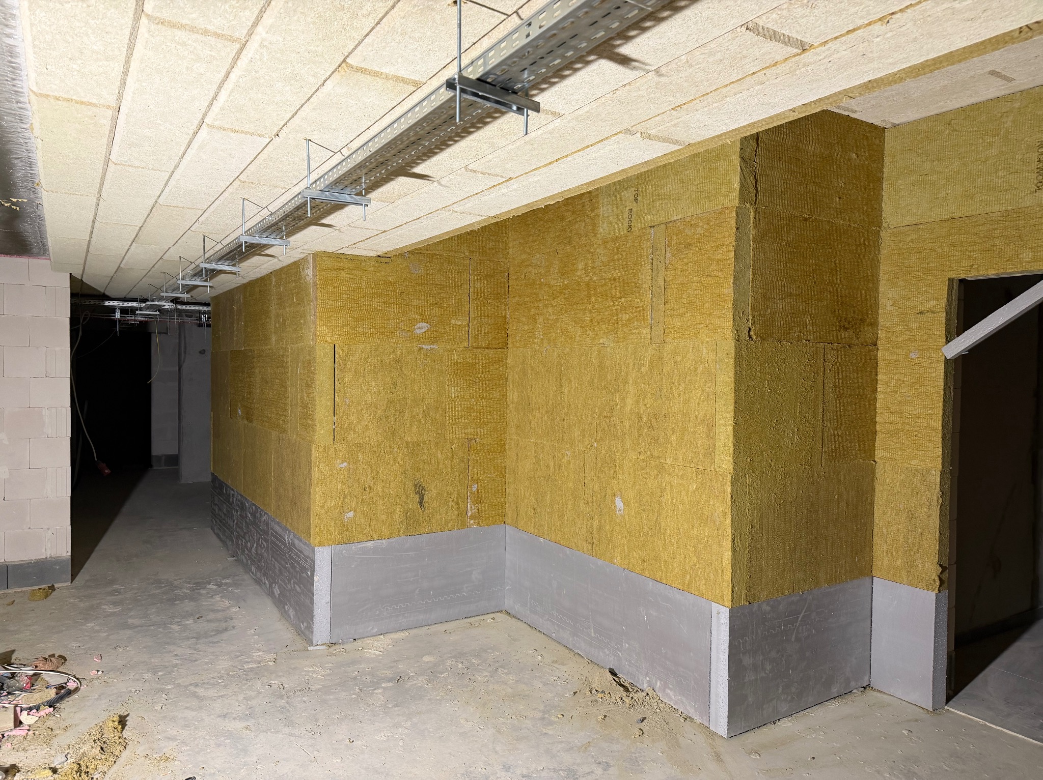 Ocieplone ściany wełną mineralną w surowym wnętrzu. Widoczne szyny pod sufitem i fragment ściany z bloczków. Szare płyty przy podłodze. Etap prac wykończeniowych.