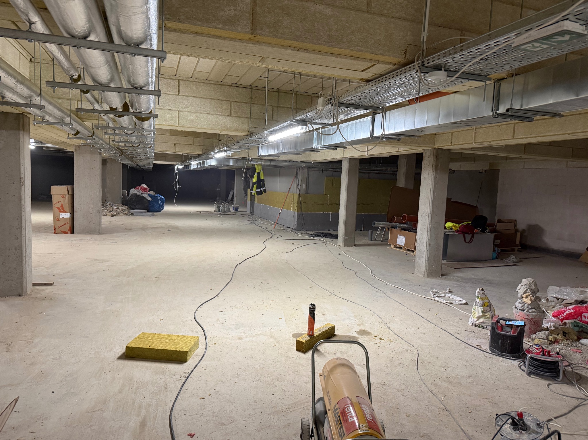 Wnętrze w trakcie prac wykończeniowych: widoczne ocieplenie stropu wełną mineralną, instalacje pod sufitem, betonowe kolumny i narzędzia budowlane na posadzce.