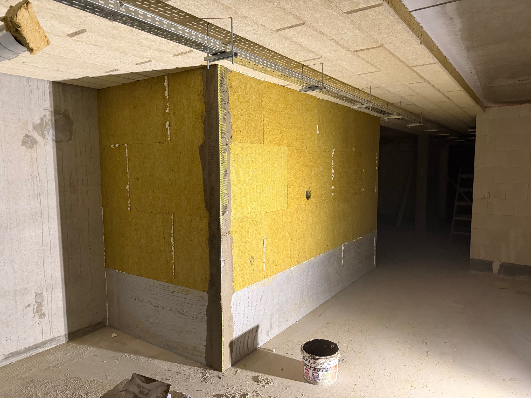 Ocieplenie ścian wełną mineralną wewnątrz budynku, widoczne profile pod instalacje elektryczne pod sufitem, grunt w wiaderku na podłodze. Etap prac wykończeniowych.