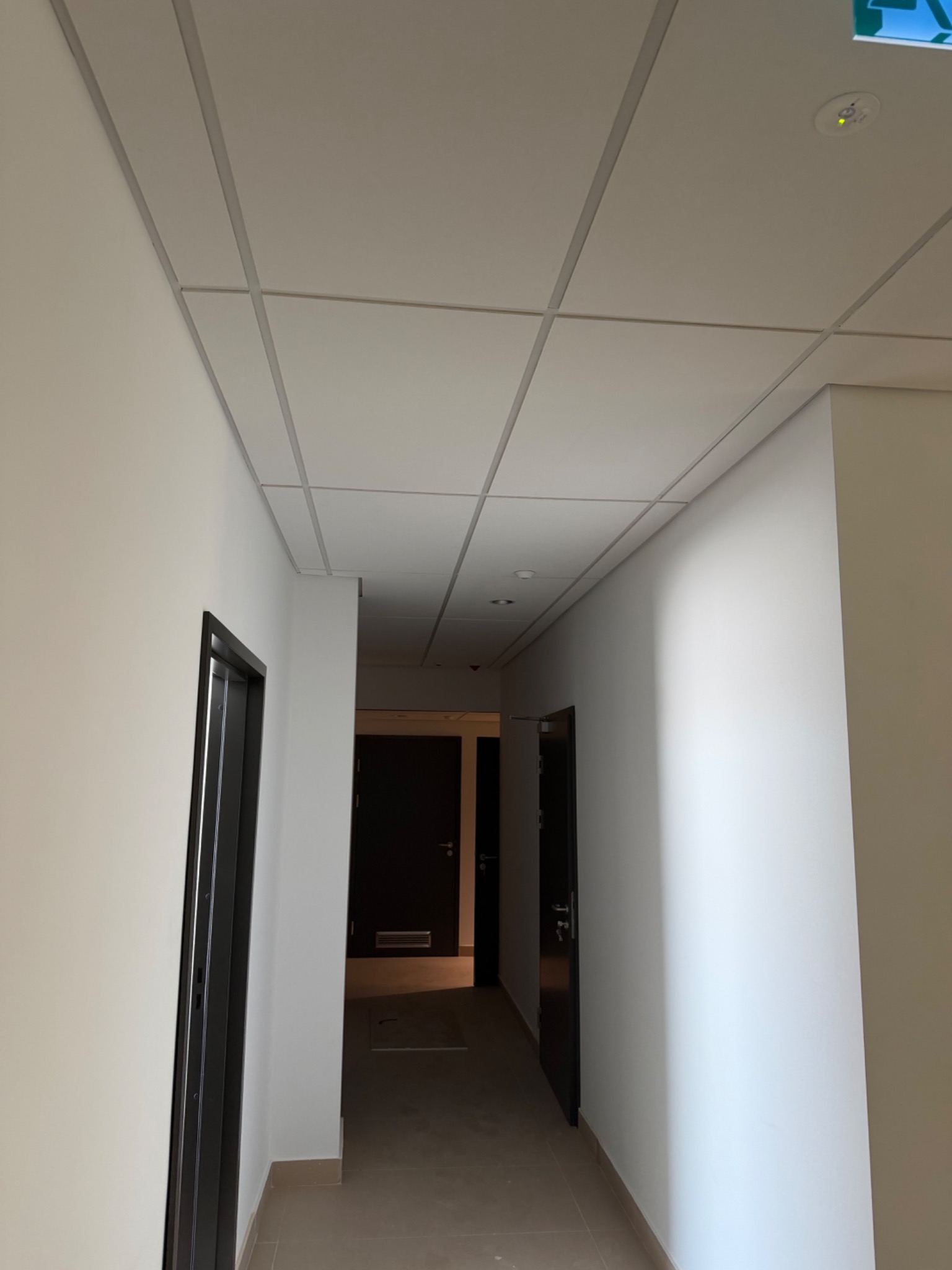Korytarz biurowy z podwieszanym sufitem kasetonowym i świeżo pomalowanymi ścianami na biało. Widoczne drzwi i oświetlenie.