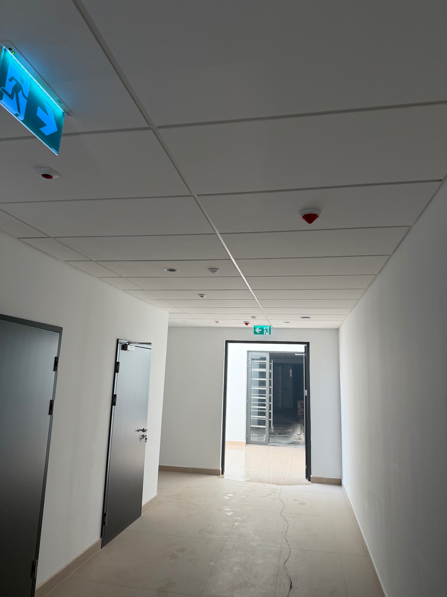 Korytarz biurowy z podwieszanym sufitem rastrowym i znakami ewakuacyjnymi. Widoczne drzwi i fragment posadzki. Świeżo po remoncie, gotowe do użytku.