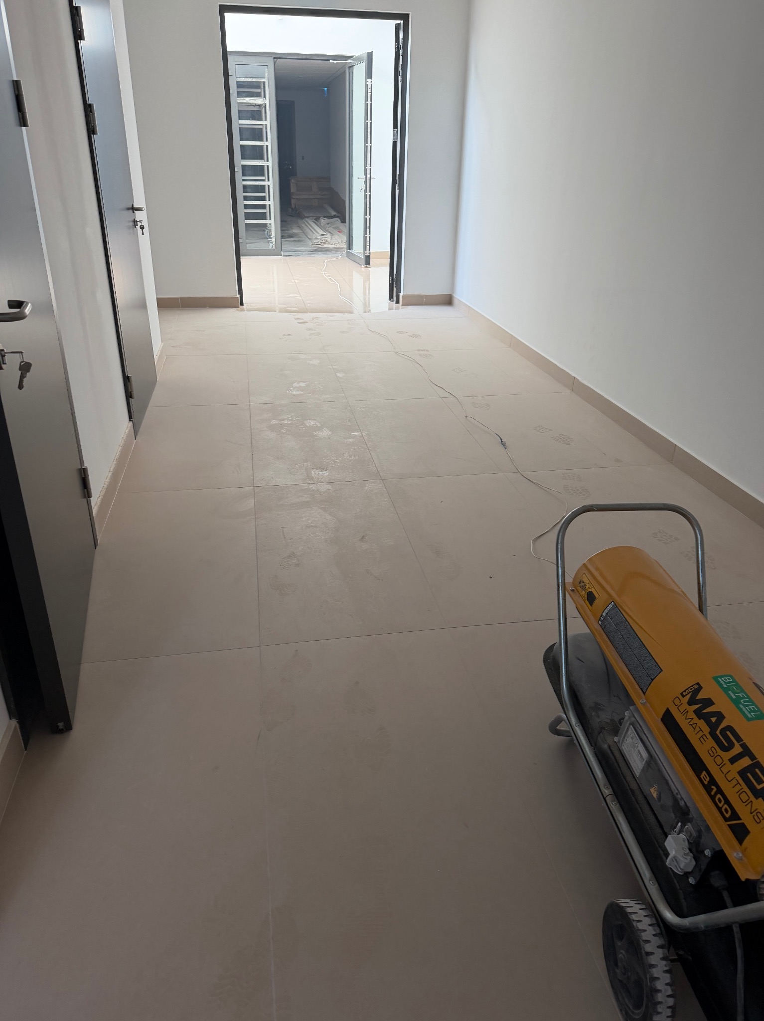 Wykończony korytarz z beżową podłogą z płytek, widoczne ślady po pracach. W tle otwarte drzwi z widokiem na dalsze pomieszczenia. Nagrzewnica Master B100 w rogu.