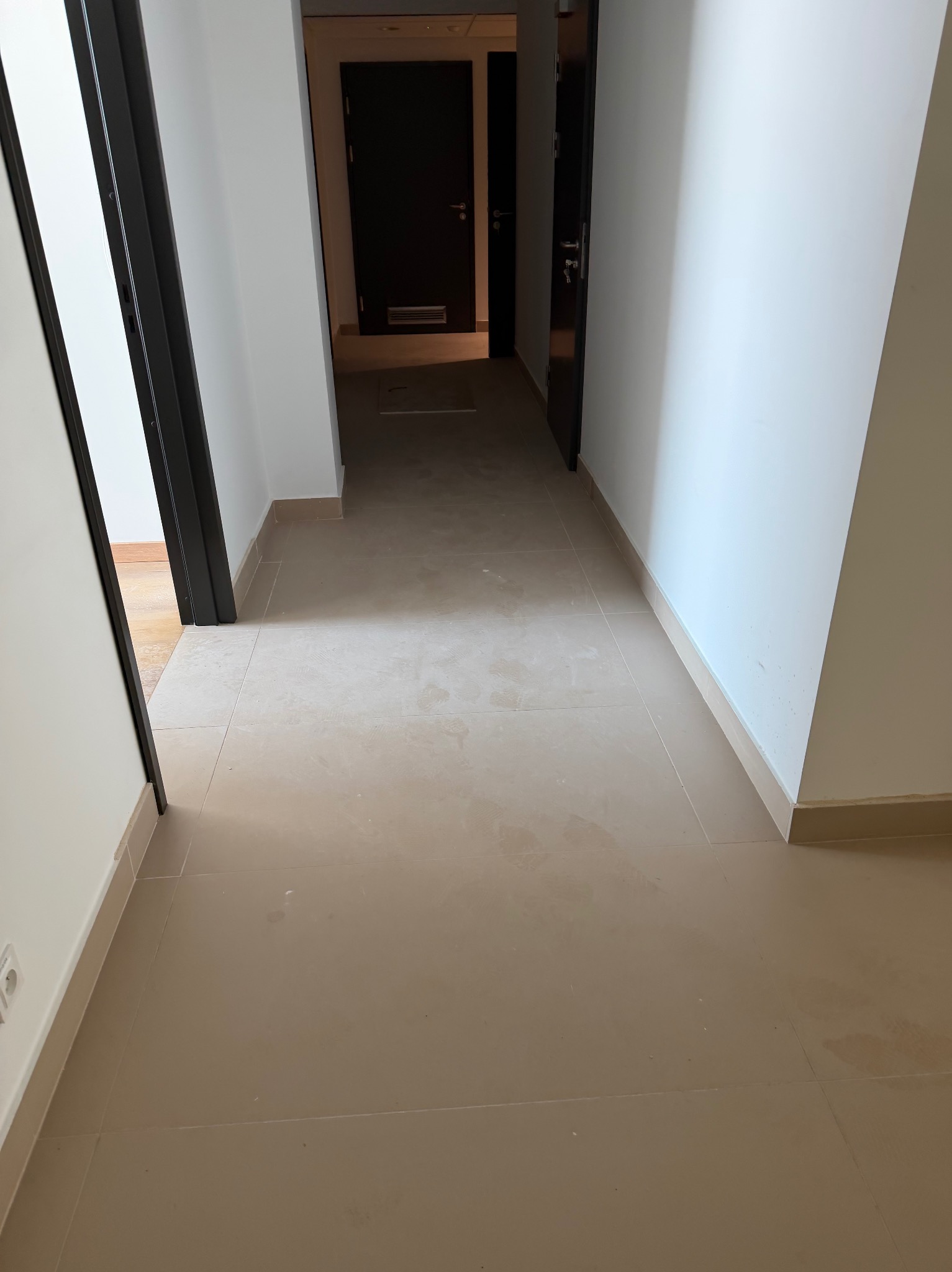 Beżowa, błyszcząca podłoga z płytek w korytarzu. Widoczne drzwi w ciemnej futrynie i białe ściany. Minimalistyczny design wnętrza.