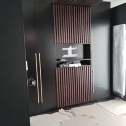 Czarna szafa z drewnianymi lamelami i złotymi uchwytami, z wnęką na telewizor, w trakcie montażu, podłoga zabezpieczona kartonem.