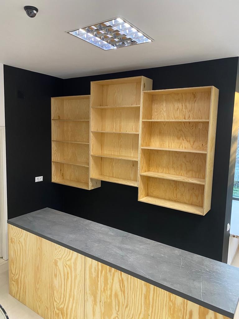 Trzy surowe, drewniane regały wiszące na czarnej ścianie nad blatem z szarego kamienia, widoczne oświetlenie sufitowe i kamera przemysłowa.