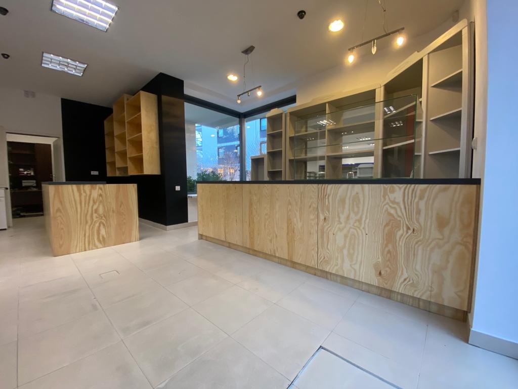 Wnętrze lokalu usługowego z zabudową meblową z surowej sklejki: regały sklepowe z przeszkloną górną częścią i niskie szafki z blatem, podłoga wyłożona jasnymi płytkami.