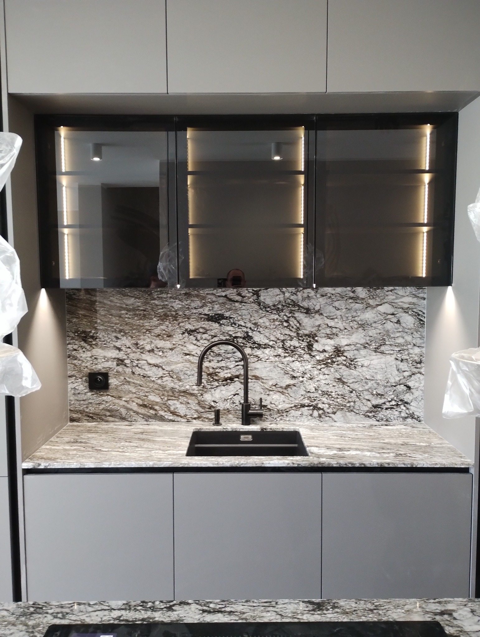 Minimalistyczna zabudowa kuchenna z szarymi frontami, czarnym zlewem i baterią oraz blatem i ścianą nad blatem z marmurkowym wzorem. Górne szafki przeszklone z podświetleniem LED.