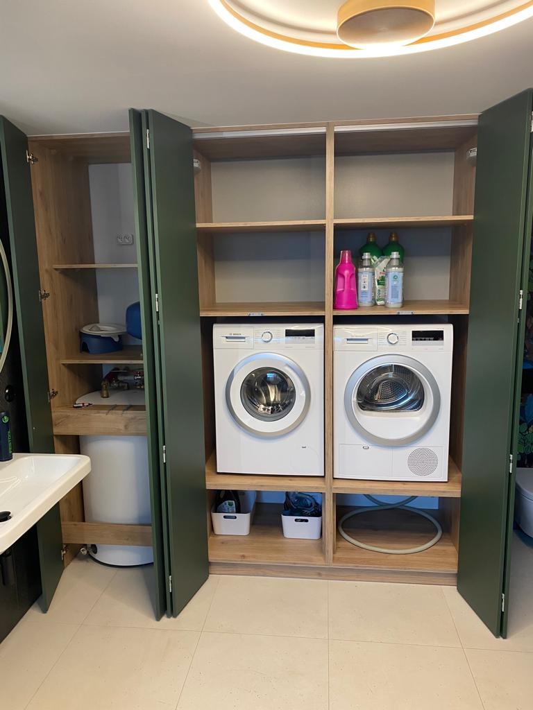 Pralka i suszarka w zabudowie z zielonymi frontami, widoczny bojler i umywalka w pomieszczeniu gospodarczym.