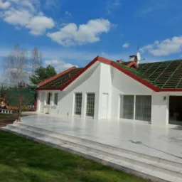 Biały dom z czerwonymi obróbkami blacharskimi i częściowo zdjętym starym pokryciem, widok z poziomu trawnika w słoneczny dzień.