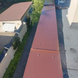 Montaż brązowej obróbki blacharskiej na dachu pokrytym papą, widok z góry, w tle budynek z ogrodem.