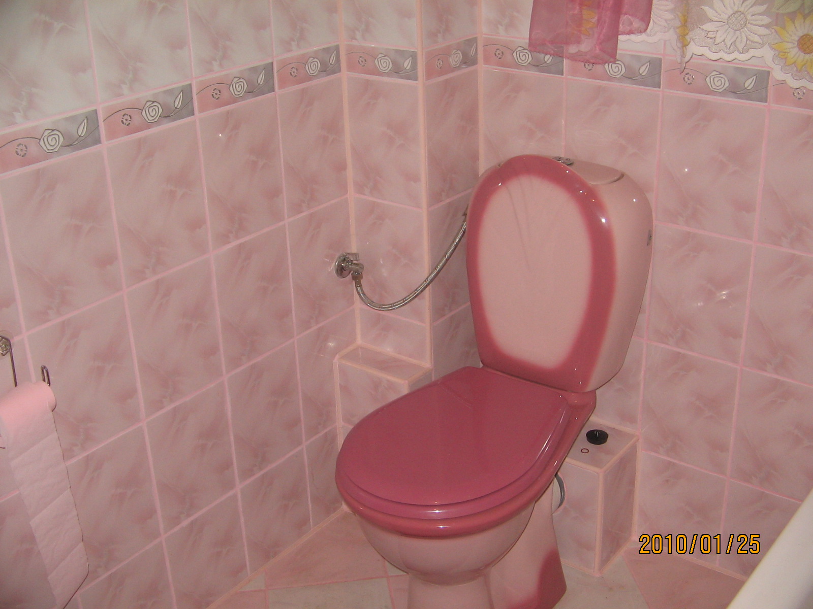 Różowa toaleta w łazience wyłożonej różowymi płytkami z dekoracyjnym pasem. Widoczny zawór doprowadzający wodę i uchwyt na papier toaletowy z różowym papierem.