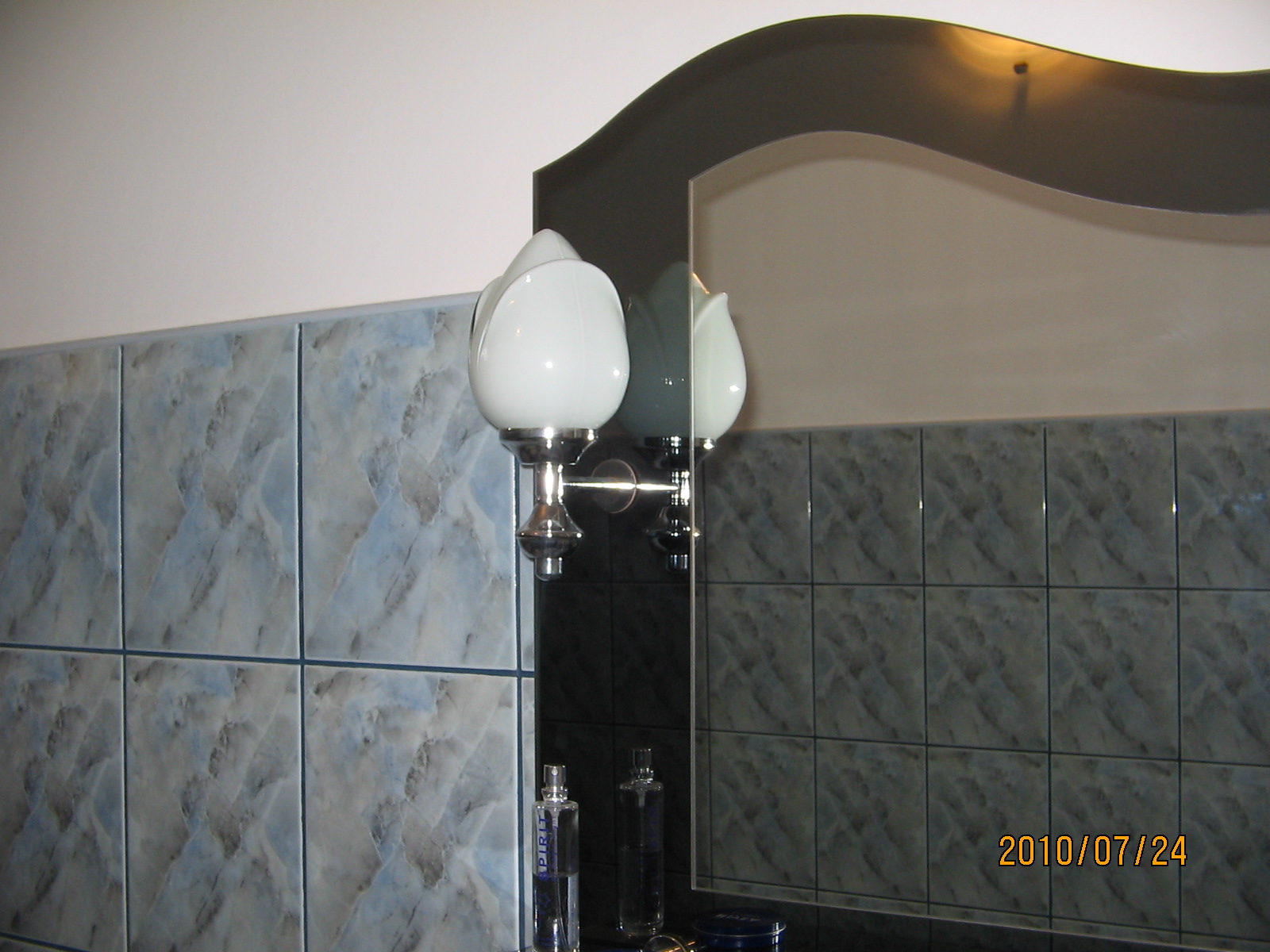 Łazienka z niebiesko-szarymi płytkami imitującymi marmur, ozdobnym lustrem z ciemną ramą i lampą w stylu retro z mleczną, tulipanową kulą.