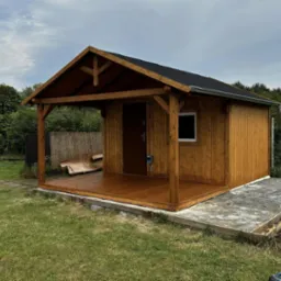 Nowoczesna, drewniana altana z zadaszonym tarasem, wykończona ciemnym drewnem, na tle zielonego ogrodu i pochmurnego nieba.