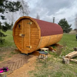 Drewniana sauna w kształcie beczki z czerwonym dachem z gontów, umieszczona na trawniku z drewnianym chodnikiem i stolikiem obok.