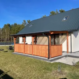 Nowoczesny dom z drewnianym tarasem i balustradą, widok z trawnika w słoneczny dzień. Widoczne czarne krzesła i stół ogrodowy. Część tarasu wyłożona kostką brukową.