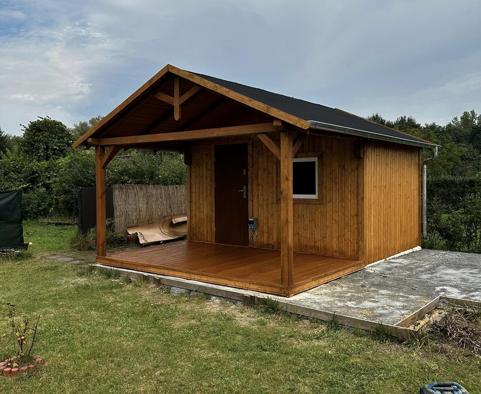 Nowoczesna, drewniana altana z zadaszonym tarasem, wykończona ciemnym drewnem, na tle zielonego ogrodu i pochmurnego nieba.