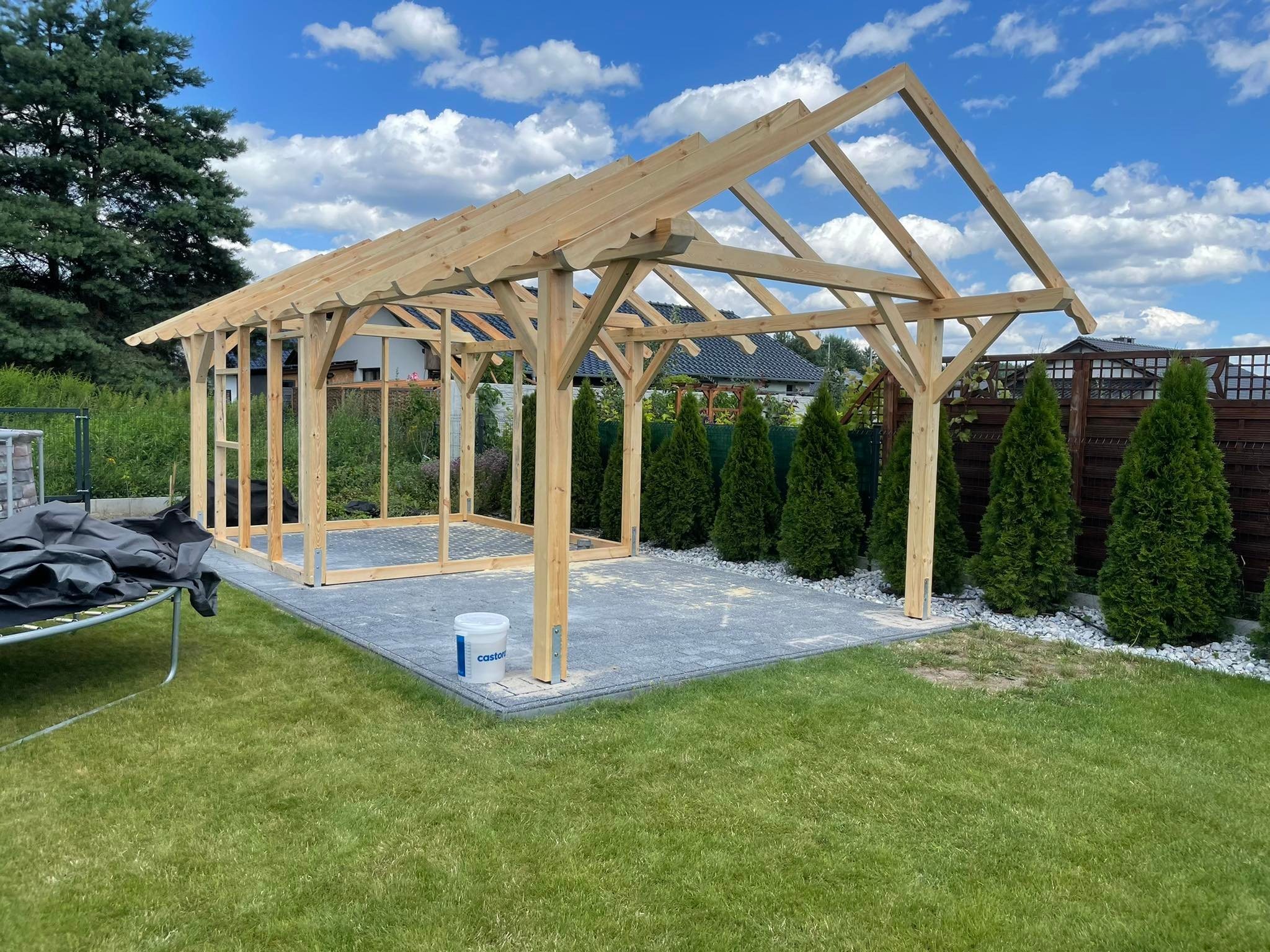 Drewniana konstrukcja altany w trakcie budowy, widoczna na tle zielonego trawnika i błękitnego nieba z chmurami. Na pierwszym planie kostka brukowa i wiadro z farbą.