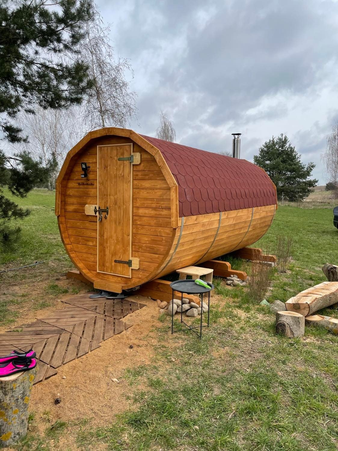 Drewniana sauna w kształcie beczki z czerwonym dachem z gontów, umieszczona na trawniku z drewnianym chodnikiem i stolikiem obok.