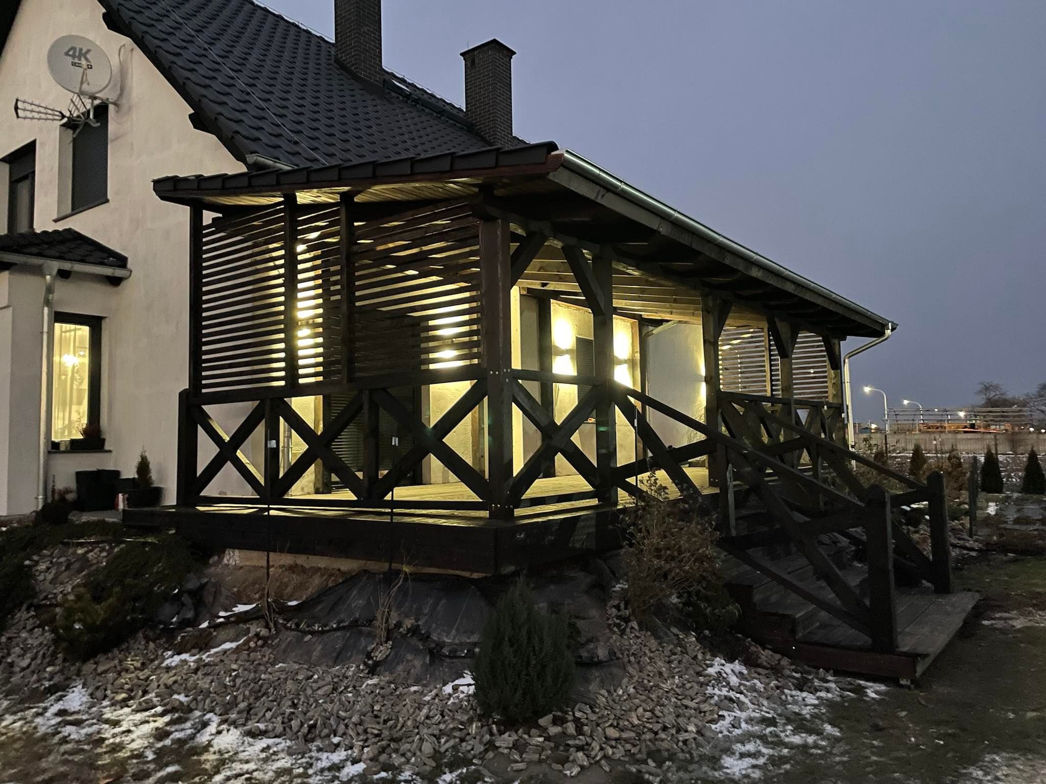 Zadaszona drewniana konstrukcja z lamelami i balustradą krzyżową, oświetlona od wewnątrz, przylegająca do domu z anteną satelitarną, widoczna w szarym świetle zmierzchu.
