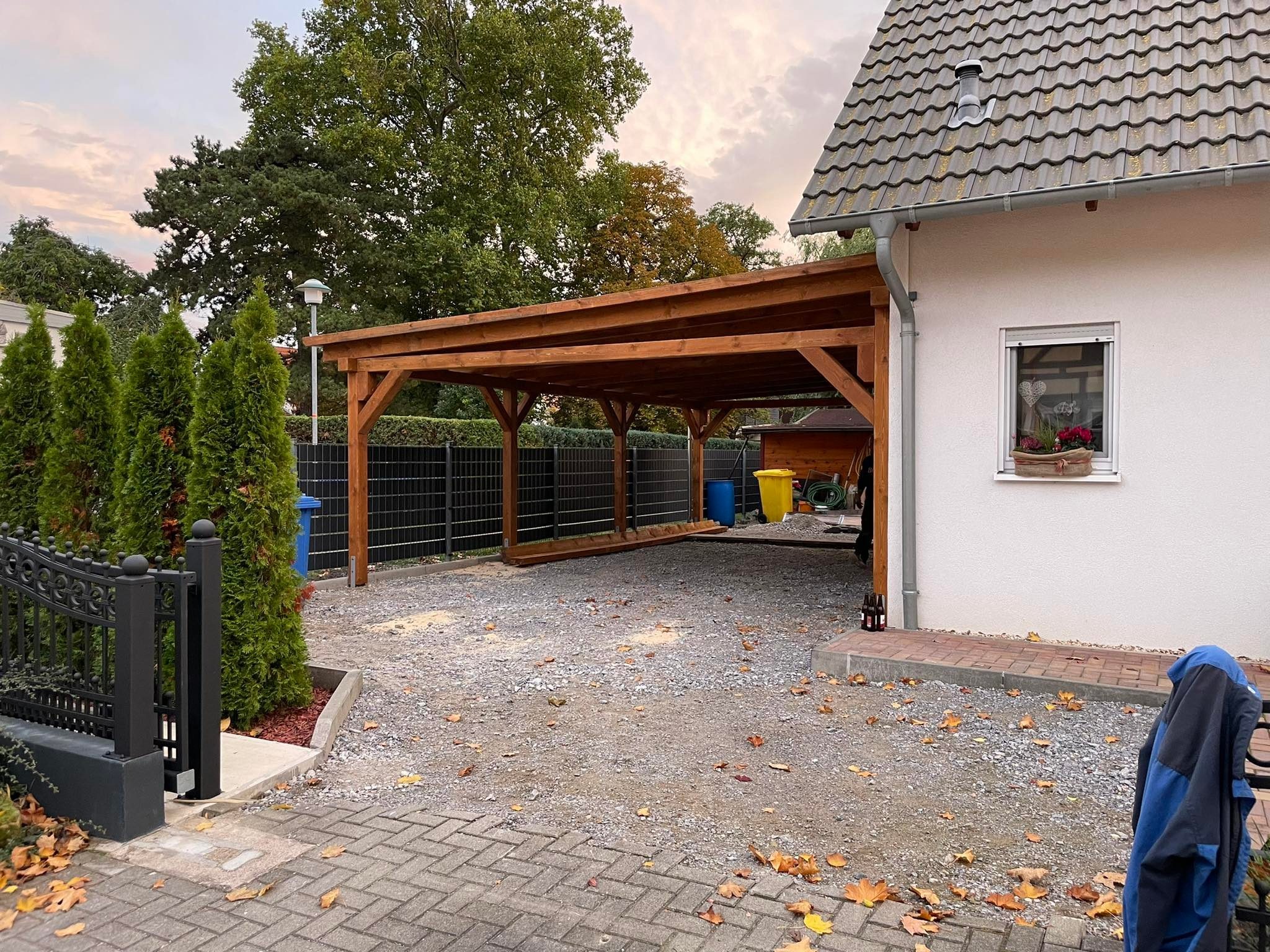 Drewniana wiata garażowa z belek na posesji z kostki brukowej, widok na dom z oknem i dekoracjami, obok tuje i brama.