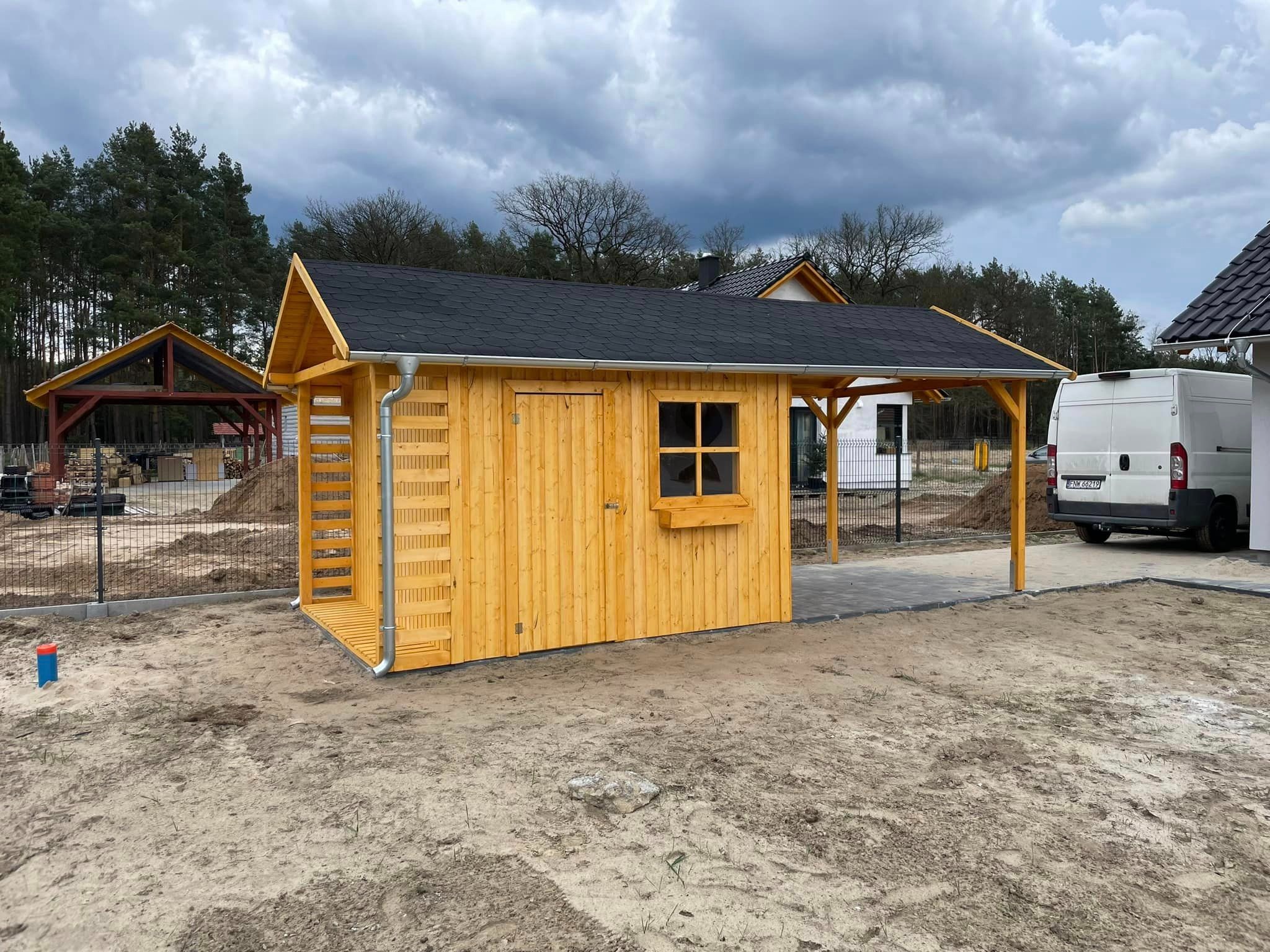 Nowa, jasna, drewniana konstrukcja z czarnym dachem, przypominająca domek narzędziowy, z rynną, małym oknem i zadaszeniem, ustawiona na piaszczystym terenie, w tle widoczny częściowo drewniany...