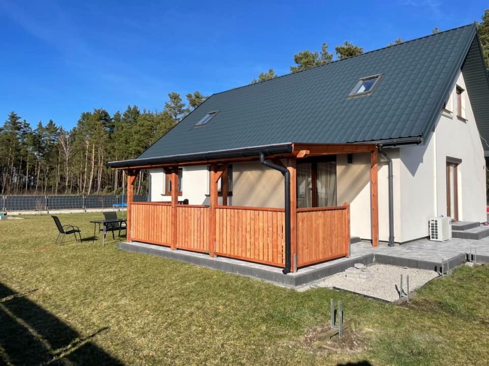 Nowoczesny dom z drewnianym tarasem i balustradą, widok z trawnika w słoneczny dzień. Widoczne czarne krzesła i stół ogrodowy. Część tarasu wyłożona kostką brukową.