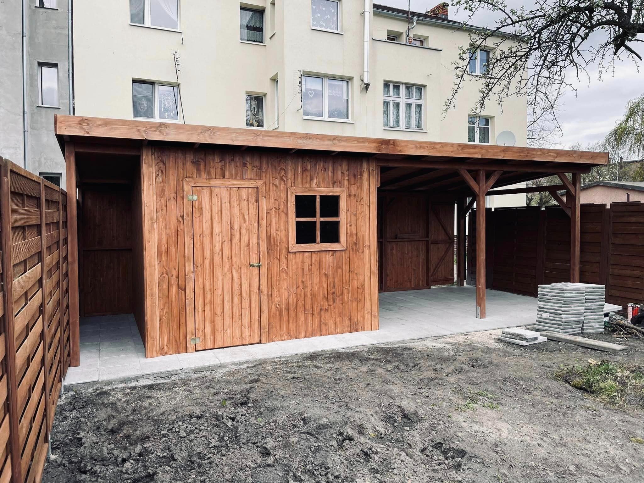 Drewniana konstrukcja z zamykanym pomieszczeniem i zadaszeniem, na betonowej wylewce, obok stos kostki brukowej, w tle budynek mieszkalny.