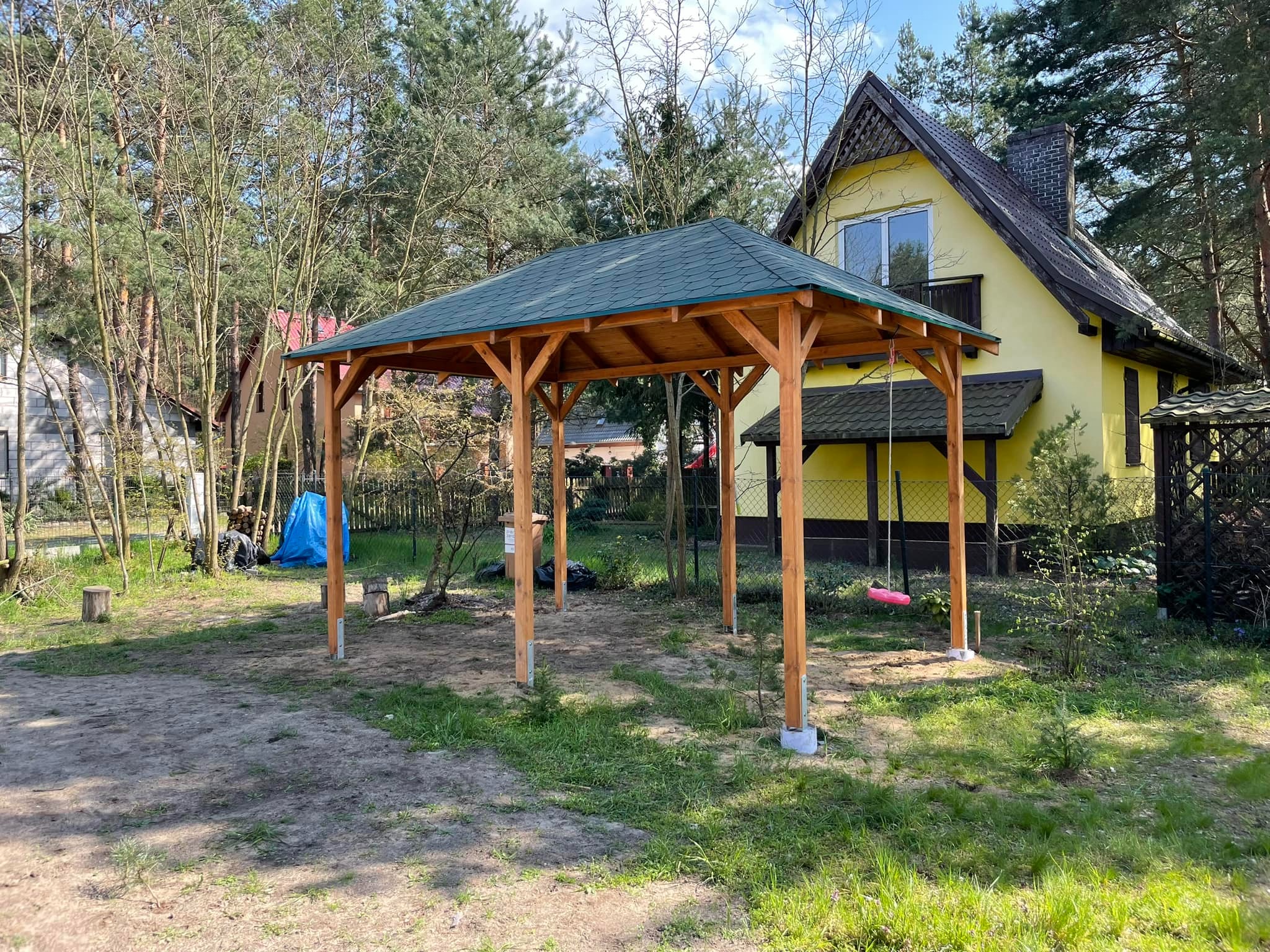 Nowo postawiona drewniana konstrukcja altany z zielonym dachem na działce z widocznym domem w tle i huśtawką.
